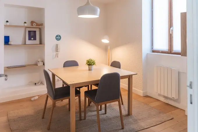 Bureau de taille moyenne carrée avec une fenêtre, une table carrée avec quatres chaises à chaque côté sur un tapis, lampes, plantes et étagères dans la pièce.