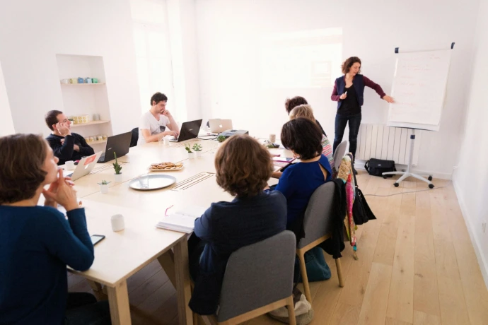 Personnes en réunion dans la salle de formation, une personne au paper board et les autres assis sur leur ordinateur.