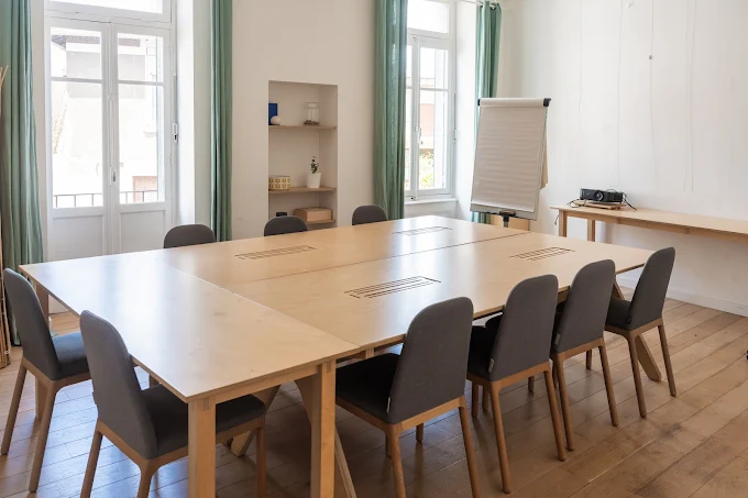 Salle de formation, large pièces avec deux grandes fenêtres. Grande table en bois au milieu avec une dizaine de chaises autour, tableau paper board et projecteur