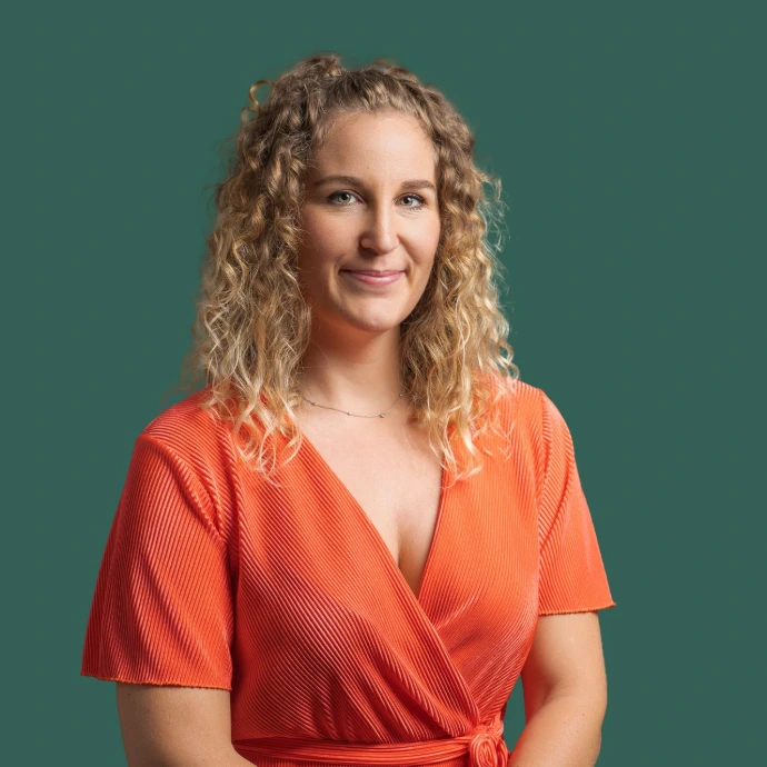 Buste de Fanny en haut rouge à manche mi courtes et fond bleu foncé, fanny se tient droite avec un sourire, les cheveux bruns à la racine et blonds au bout, cheveux mi long